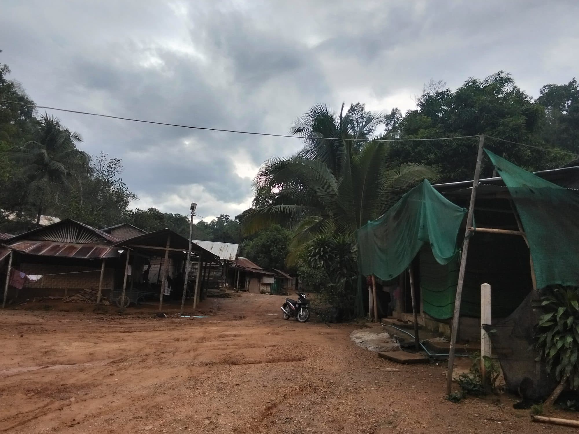 A dirt road running through the off-grid village, past tin-roofed and bamboo homes under a monsoon sky.